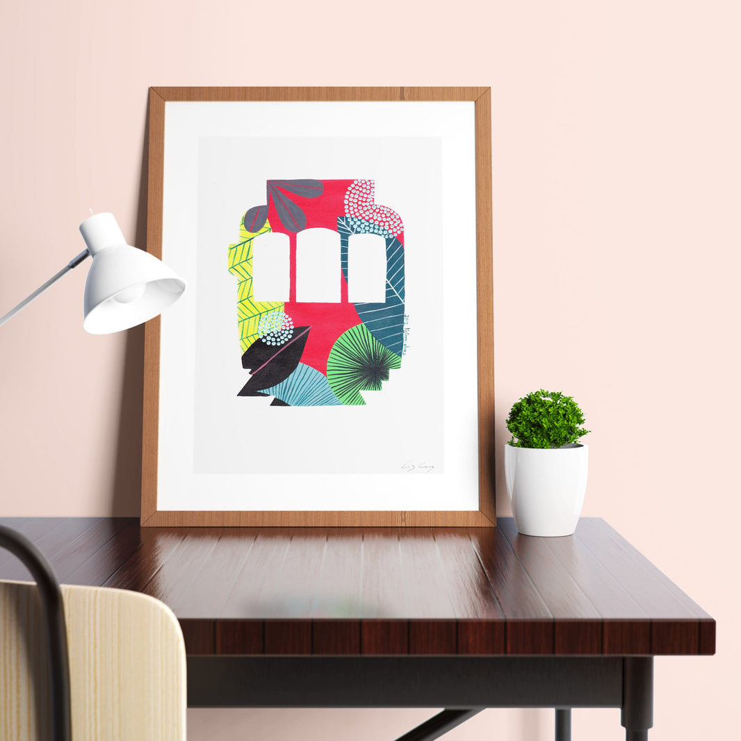 A framed fine art print, by Lis Gonçalves. Open edition, signed by the artist (printed in Hahnemühle matt fibre paper) featuring a colorful Lisbon tram displayed on a desk with a lamp and plant.