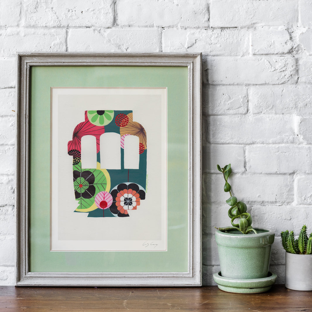 A framed fine art print, by Lis Gonçalves. Open edition, signed by the artist (printed in Hahnemühle matt fibre paper) featuring a colorful Lisbon tram on a white brick wall with potted plants.