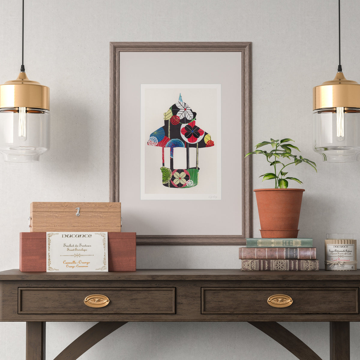 A framed fine art print, by Lis Gonçalves. Open edition, signed by the artist (printed in Hahnemühle matt fibre paper)  featuring a colorful, stylized Lisbon Kiosk, displayed on a dark wood console table in a styled room setting with two hanging pendant lights, a potted plant, and decorative boxes.