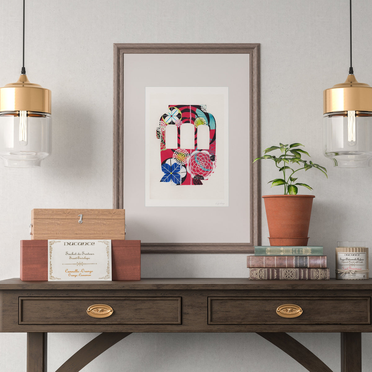A framed fine art print, by Lis Gonçalves. Open edition, signed by the artist (printed in Hahnemühle matt fibre paper) featuring a colorful Lisbon tram hangs on a light gray wall above a dark wood console table. The scene is styled with two gold and clear pendant lights, a small potted plant, books, and decorative boxes.