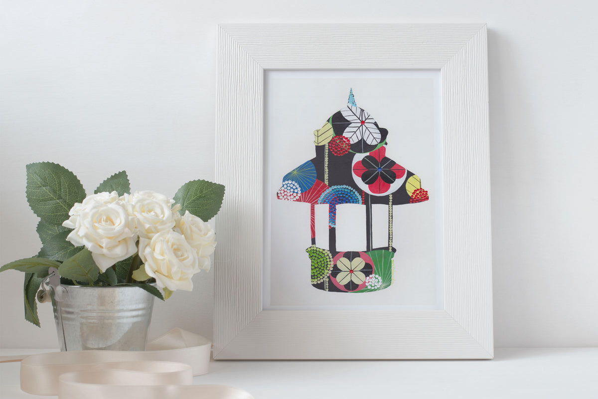 A framed fine art print, by Lis Gonçalves. Open edition, signed by the artist (printed in Hahnemühle matt fibre paper) of a colorful Lisbon kiosk on a white surface with flowers and candles.