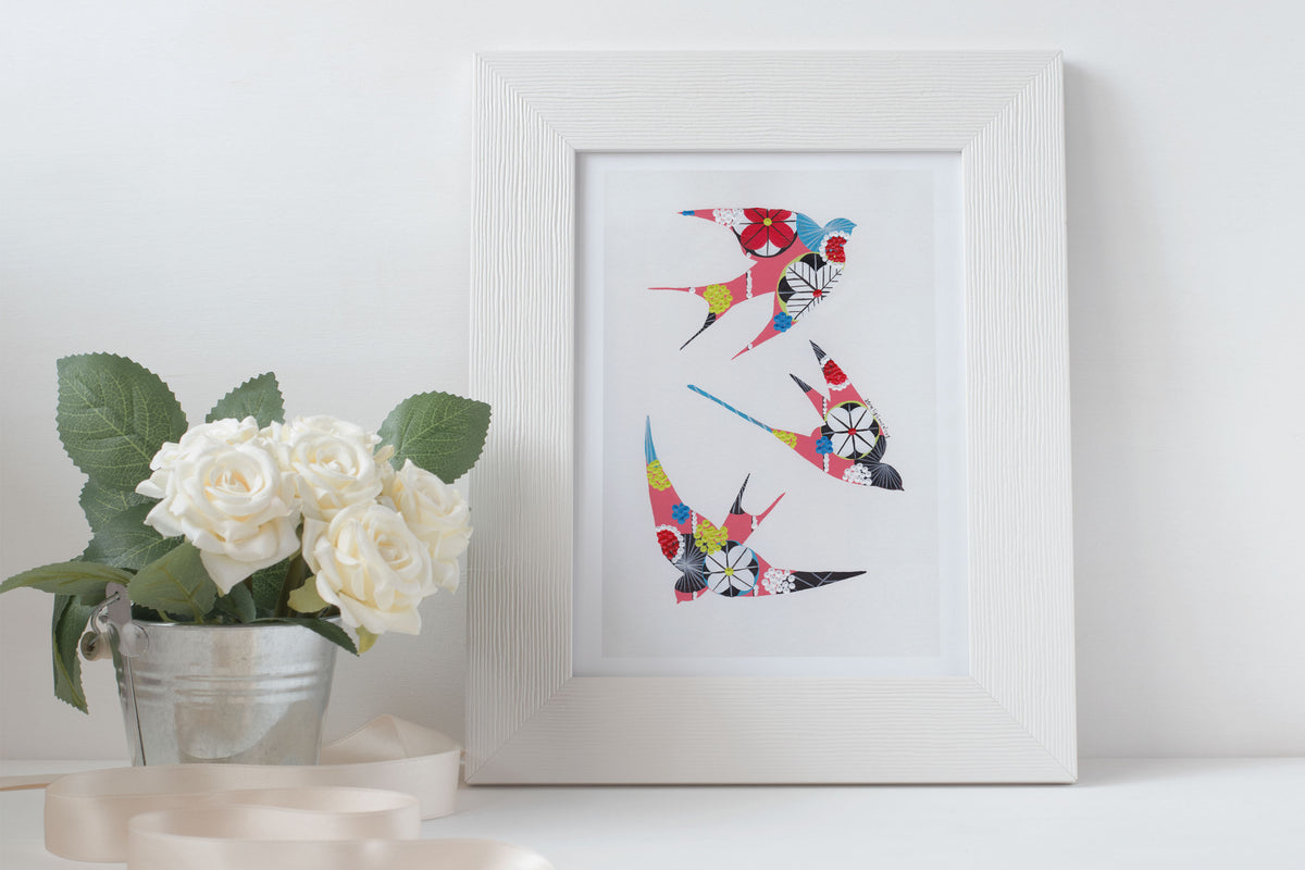 A framed fine art print, by Lis Gonçalves. Open edition, signed by the artist (printed in Hahnemühle matt fibre paper) featuring colorful swallows next to a potted plant on a white surface