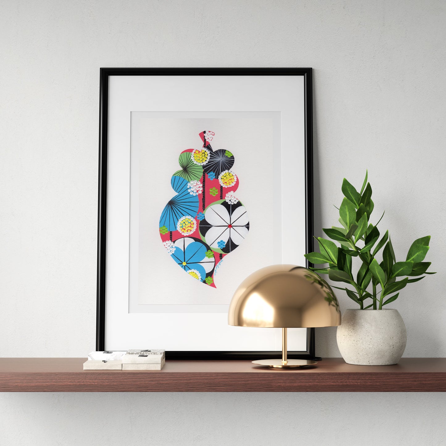 A framed fine art print, by Lis Gonçalves. Open edition, signed by the artist (printed in Hahnemühle matt fibre paper) featuring a colorful Portuguese Viana heart hangs on a light gray wall above a wooden shelf. The shelf is styled with a gold dome lamp and a potted green plant.
