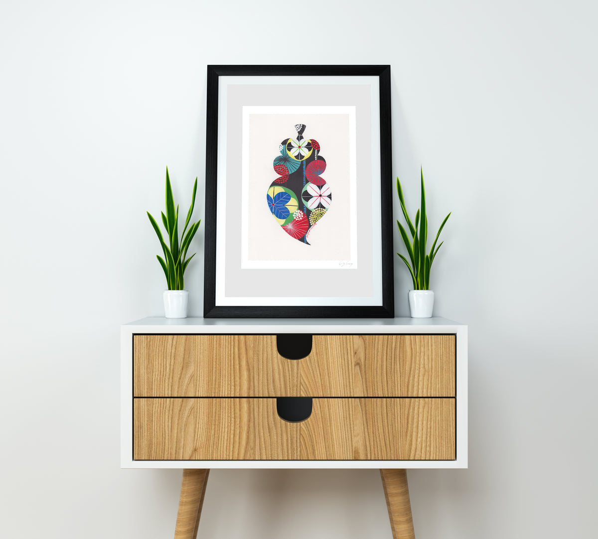A framed fine art print, by Lis Gonçalves. Open edition, signed by the artist (printed in Hahnemühle matt fibre paper) featuring a colorful Portuguese Viana heart on a wooden side table with plants against a white wall.