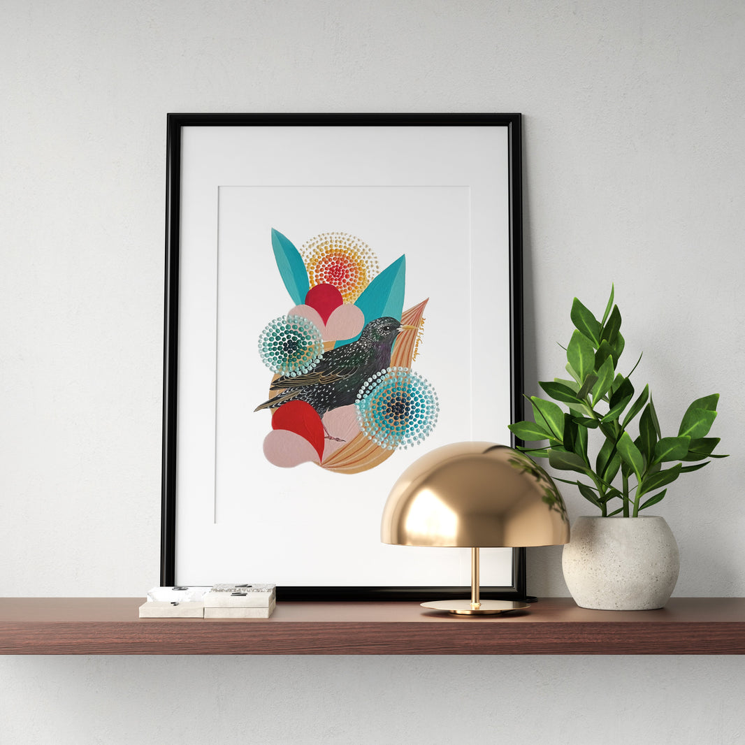 A framed fine art print, by Lis Gonçalves. Printed in Hahnemühle matt fibre paper; featuring a bird and colorful geometric shapes, displayed on a wooden shelf next to a gold dome lamp and a potted green plant against a white wall.