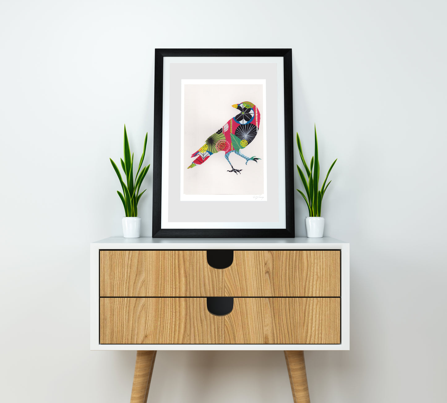 A framed fine art print, by Lis Gonçalves. Open edition, signed by the artist (printed in Hahnemühle matt fibre paper) featuring a colorful raven on a white wall above a wooden cabinet with two drawers.
