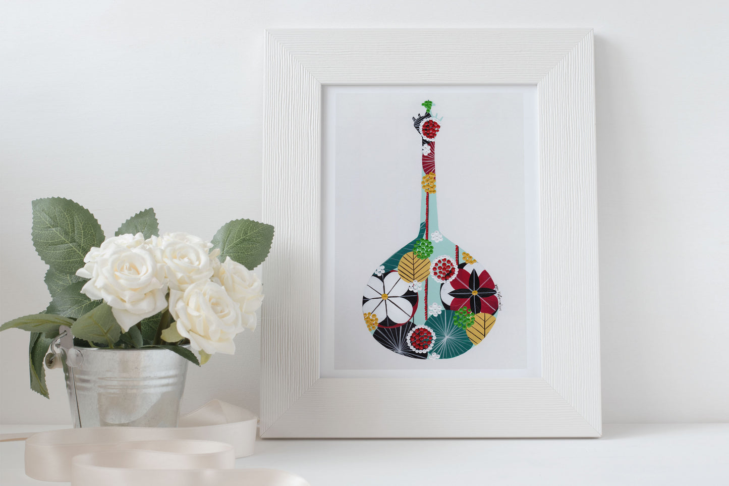 A framed fine art print, by Lis Gonçalves. Open edition, signed by the artist (printed in Hahnemühle matt fibre paper) featuring a colorful Portuguese guitar with flowers on a white surface next to a potted plant.