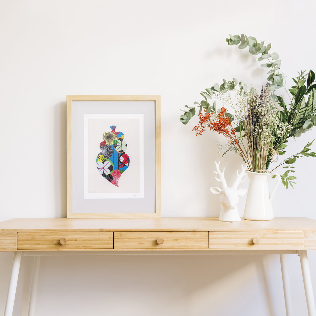 A framed fine art print, by Lis Gonçalves. Open edition, signed by the artist (printed in Hahnemühle matt fibre paper) featuring a colorful Portuguese Viana heart on a wooden desk with a vase of flowers on a white background