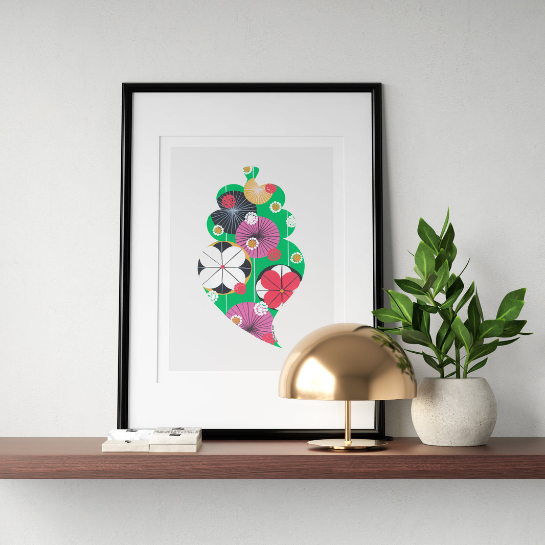 A framed fine art print, by Lis Gonçalves. Open edition, signed by the artist (printed in Hahnemühle matt fibre paper) featuring a colorful Portuguese Viana heart, styled on a wooden shelf next to a gold dome lamp and a potted green plant against a light gray wall.