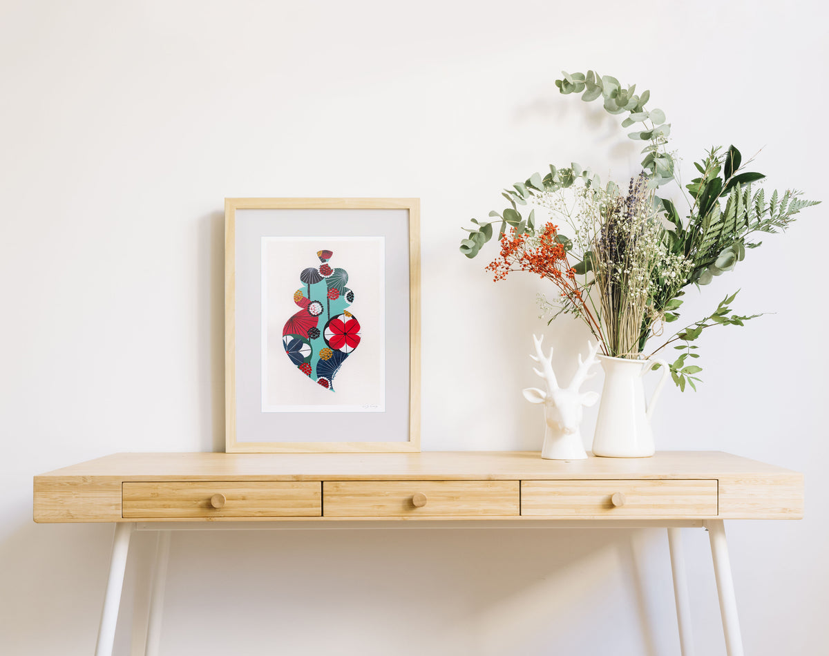 A framed fine art print, by Lis Gonçalves. Open edition, signed by the artist (printed in Hahnemühle matt fibre paper) featuring a colorful Portuguese Viana heart  on a wooden desk with a vase of flowers on a white background