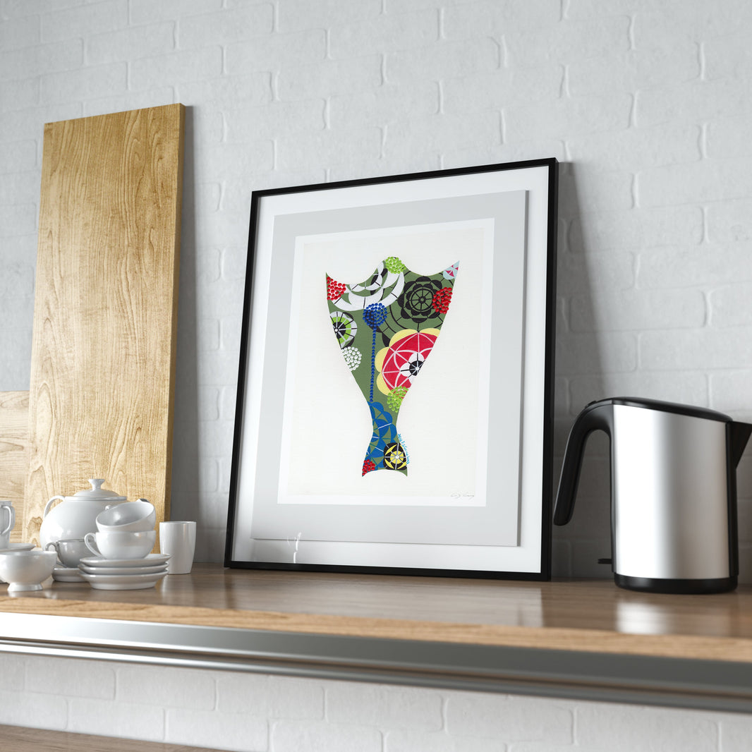 A framed fine art print, by Lis Gonçalves. Open edition, signed by the artist (printed in Hahnemühle matt fibre paper) featuring a colorful, patterned cod fish illustration in a black frame, leaning on a wooden countertop against a white brick wall next to a kettle and cutting board.