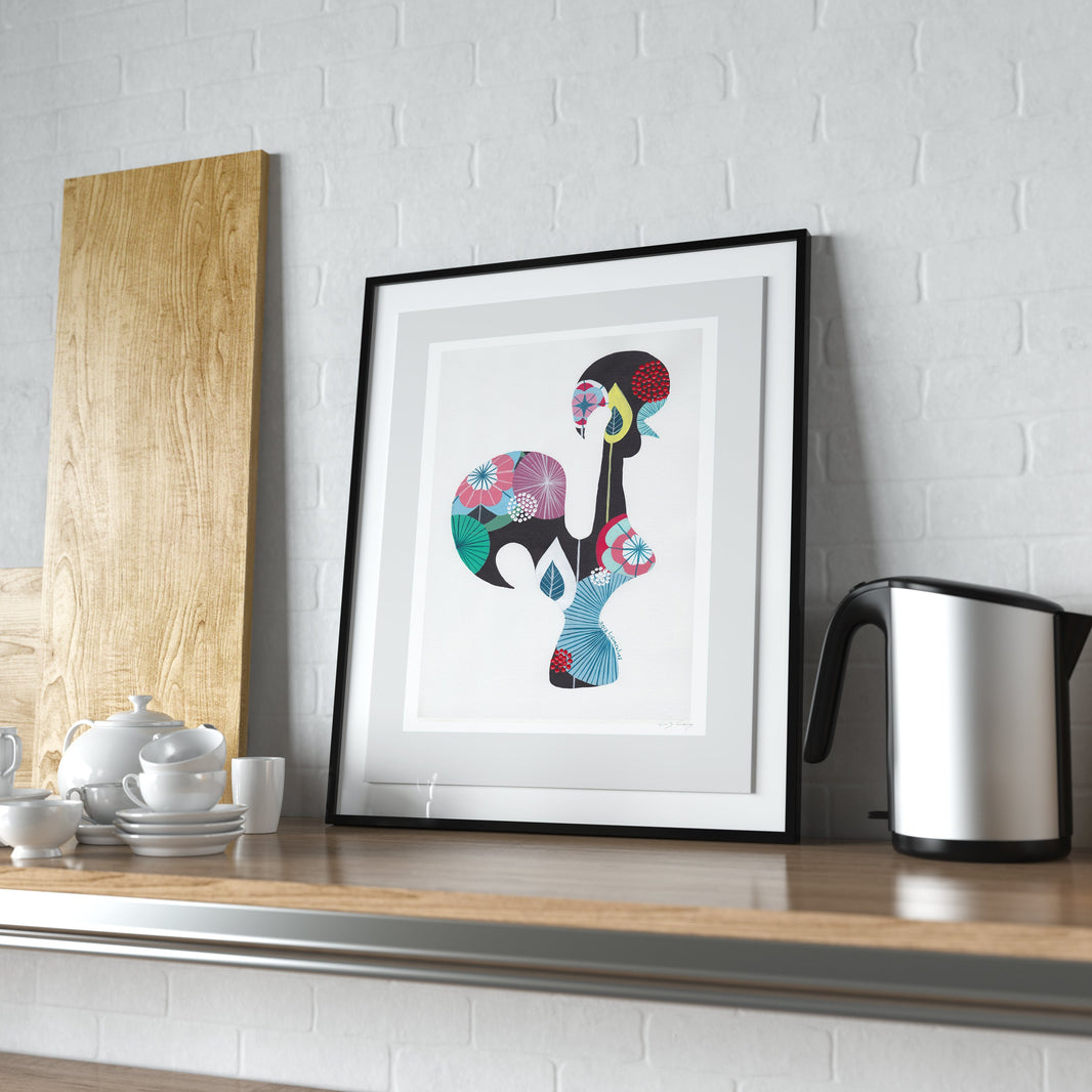 A framed fine art print, by Lis Gonçalves. Open edition, signed by the artist (printed in Hahnemühle matt fibre paper) featuring a colorful Portuguese rooster, displayed on a wooden kitchen shelf next to a kettle and teapot against a white brick wall.