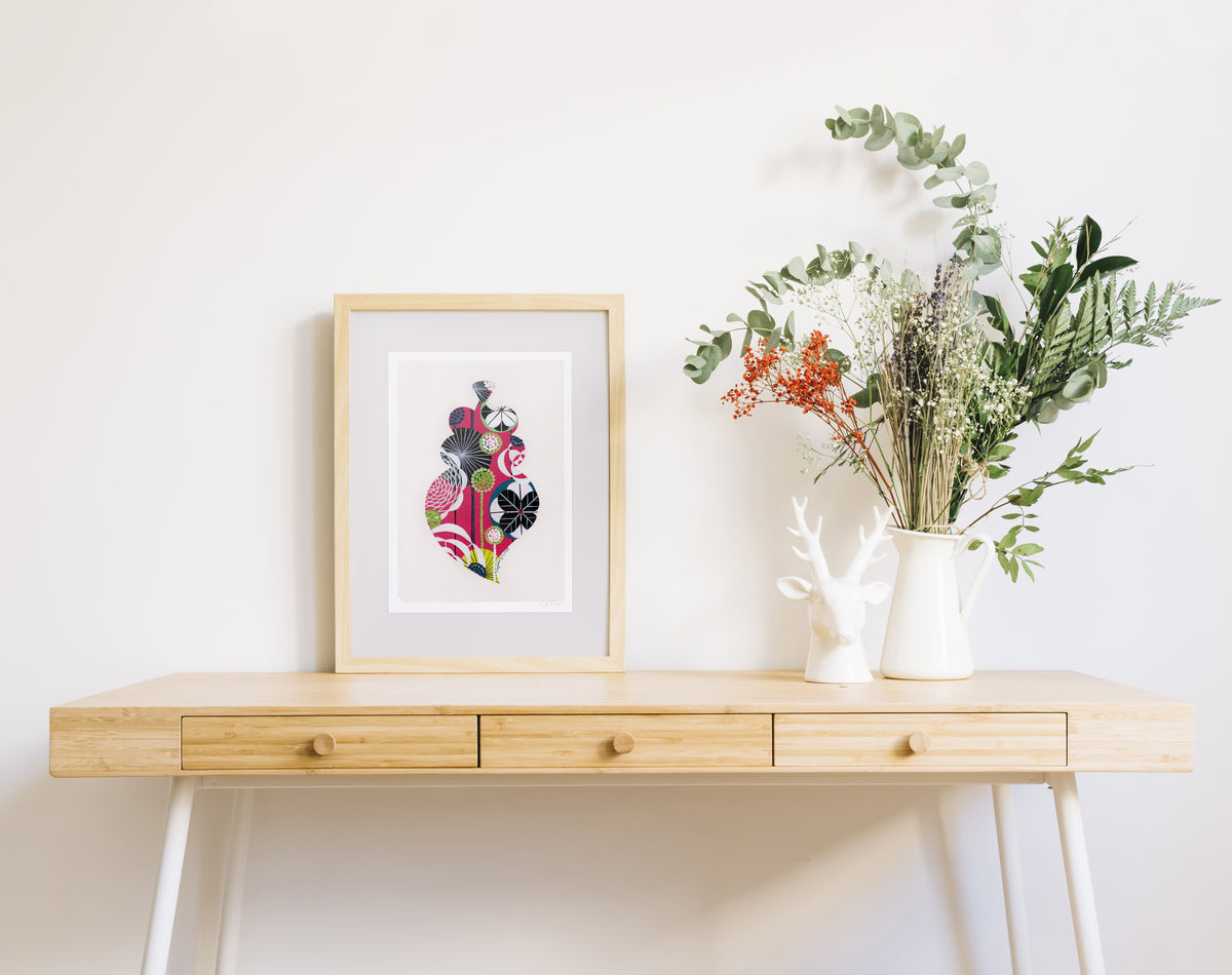 A framed fine art print, by Lis Gonçalves. Open edition, signed by the artist (printed in Hahnemühle matt fibre paper) featuring a colorful Portuguese Viana heart on a wooden desk with a vase of flowers on a white background