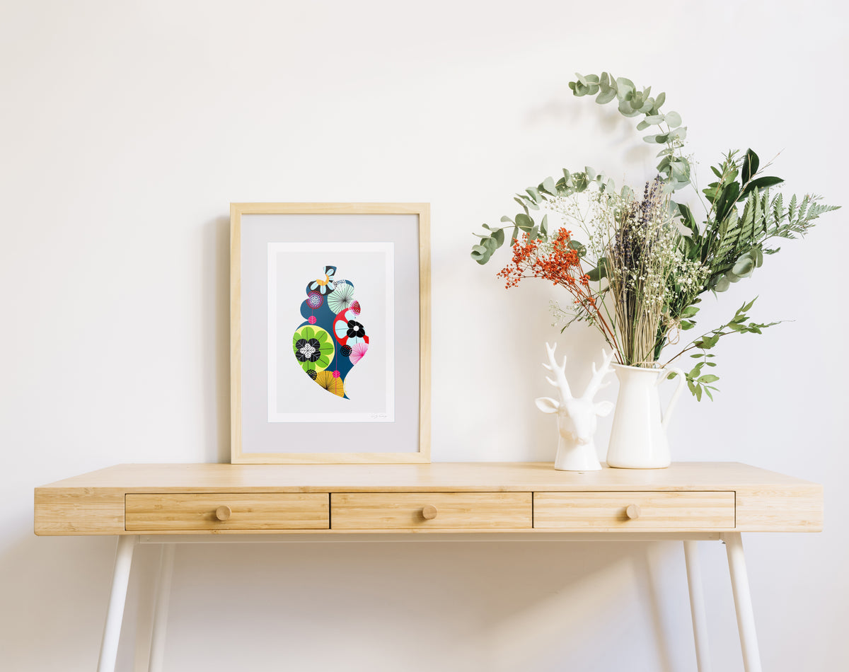 A framed fine art print, by Lis Gonçalves. Open edition, signed by the artist (printed in Hahnemühle matt fibre paper) featuring a colorful Portuguese Viana heart on a wooden desk with a vase of flowers on a white background