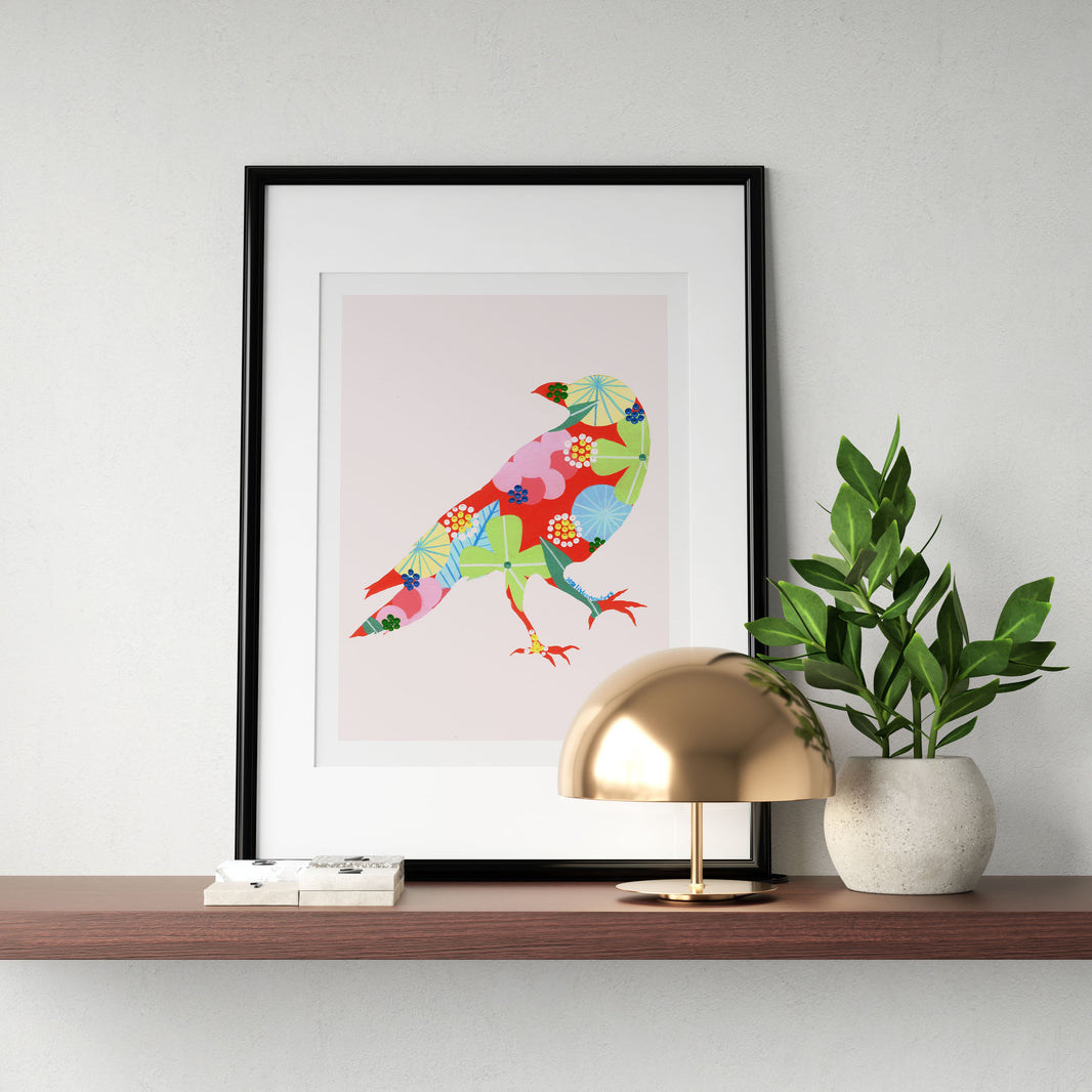 A framed fine art print, by Lis Gonçalves. Open edition, signed by the artist (printed in Hahnemühle matt fibre paper) featuring a colorful raven artwork on a wooden shelf with a lamp and plant.