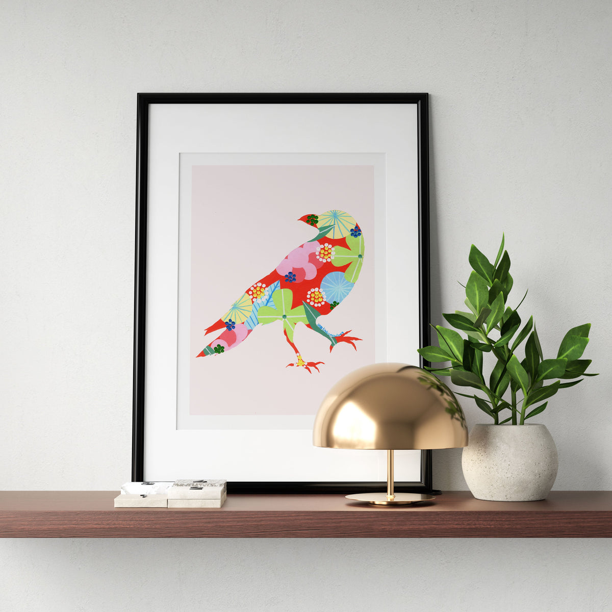 A framed fine art print, by Lis Gonçalves. Open edition, signed by the artist (printed in Hahnemühle matt fibre paper) featuring a colorful raven artwork on a wooden shelf with a lamp and plant.