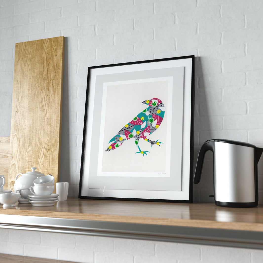 A framed fine art print, by Lis Gonçalves. Open edition, signed by the artist (printed in Hahnemühle matt fibre paper) featuring a colorful bird illustration in a black frame, styled on a wooden kitchen counter next to a kettle and a wooden board against a white brick wall.
