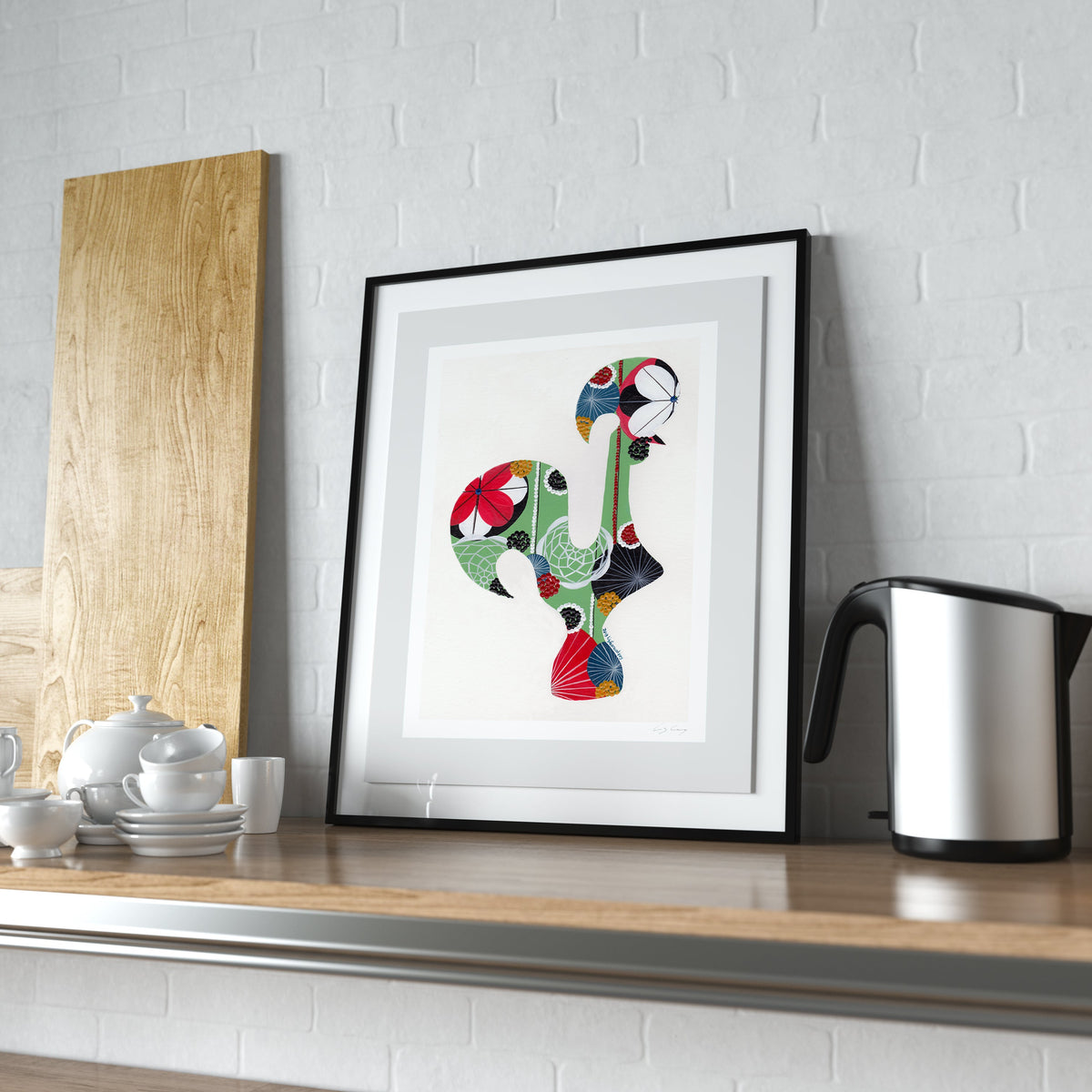A framed fine art print, by Lis Gonçalves. Open edition, signed by the artist (printed in Hahnemühle matt fibre paper) featuring a colorful Portuguese Rooster, displayed on a wooden kitchen counter next to a kettle and a wooden board against a white brick wall.
