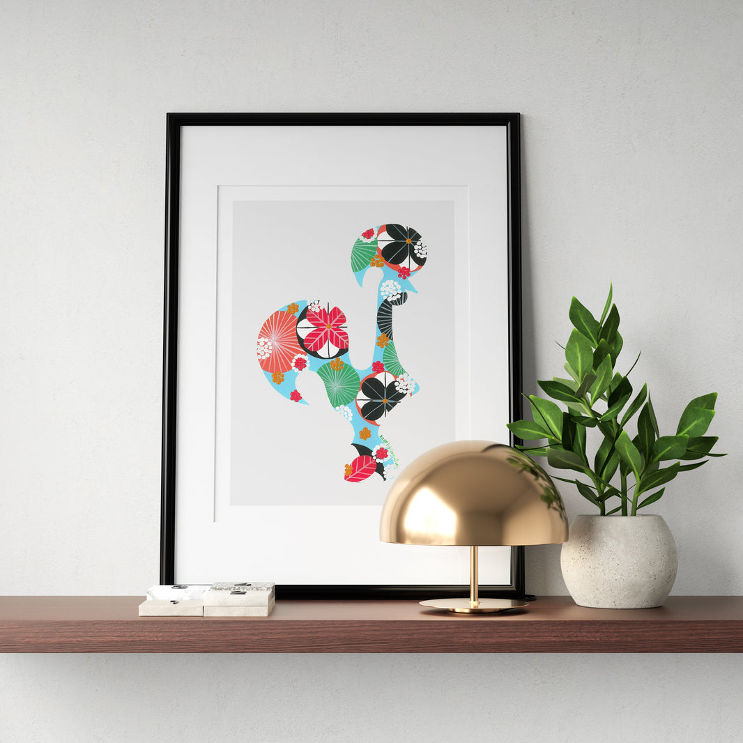 A framed fine art print, by Lis Gonçalves. Open edition, signed by the artist (printed in Hahnemühle matt fibre paper) featuring a colorful Portuguese rooster, displayed on a floating wooden shelf next to a gold dome lamp and a potted green plant against a white wall.
