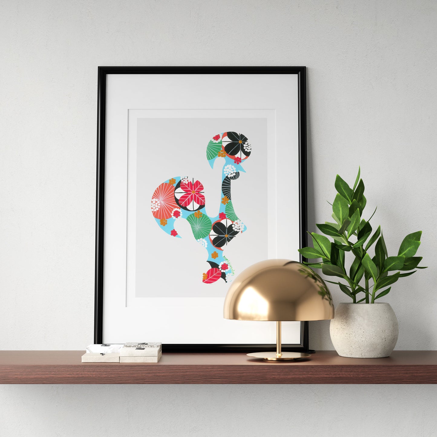 A framed fine art print, by Lis Gonçalves. Open edition, signed by the artist (printed in Hahnemühle matt fibre paper) featuring a colorful Portuguese rooster, displayed on a floating wooden shelf next to a gold dome lamp and a potted green plant against a white wall.
