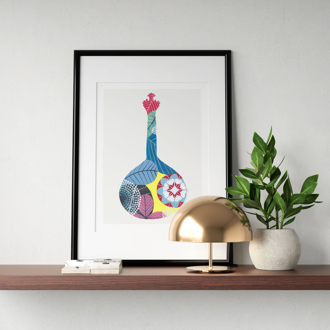 A framed fine art print, by Lis Gonçalves. Open edition, signed by the artist (printed in Hahnemühle matt fibre paper) featuring a colorful Portuguese guitar, on a wooden shelf with a lamp and plant against a white wall