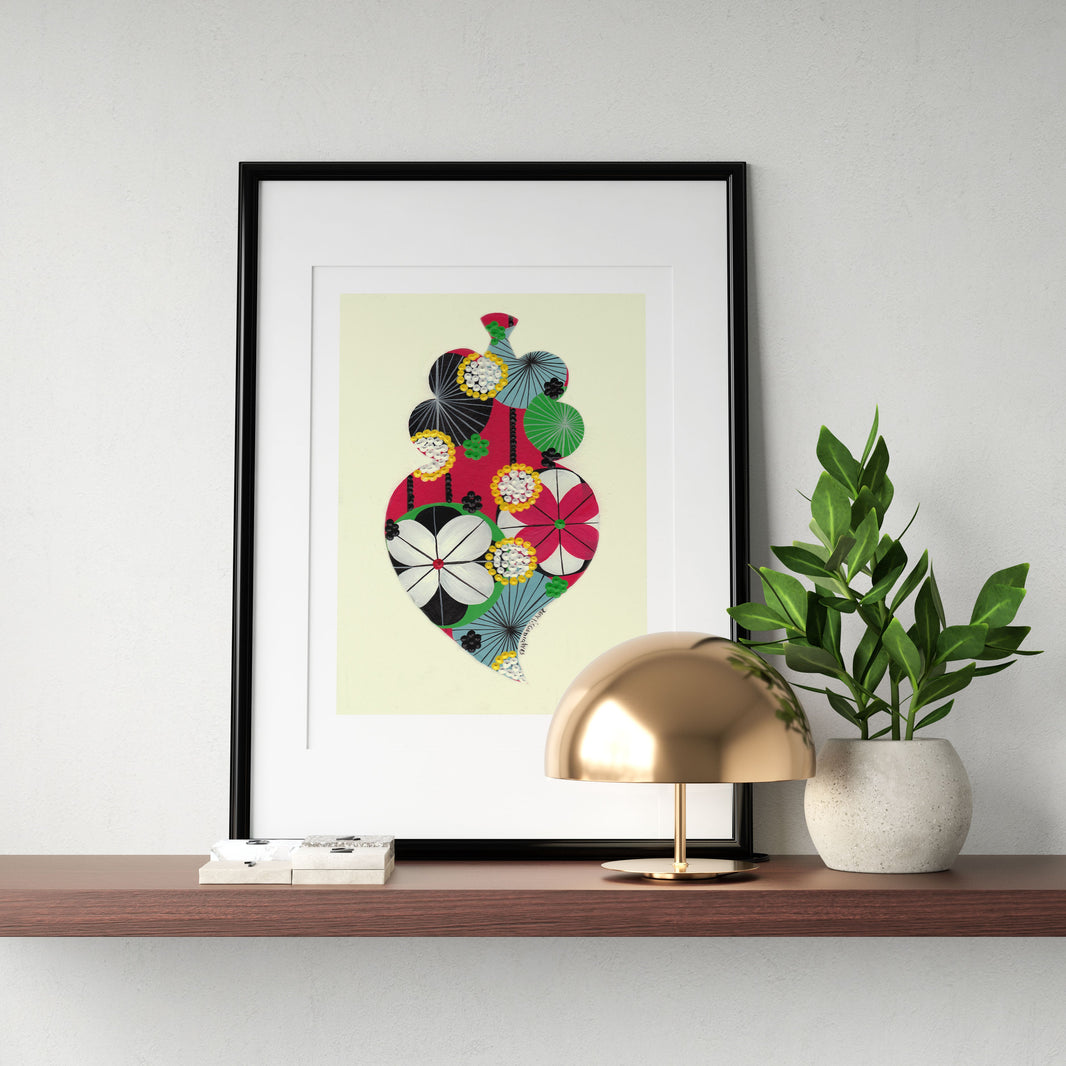 A framed fine art print, by Lis Gonçalves. Open edition, signed by the artist (printed in Hahnemühle matt fibre paper) featuring a colorful Portuguese Viana heart on a wooden shelf with a lamp and plant against a white wall.