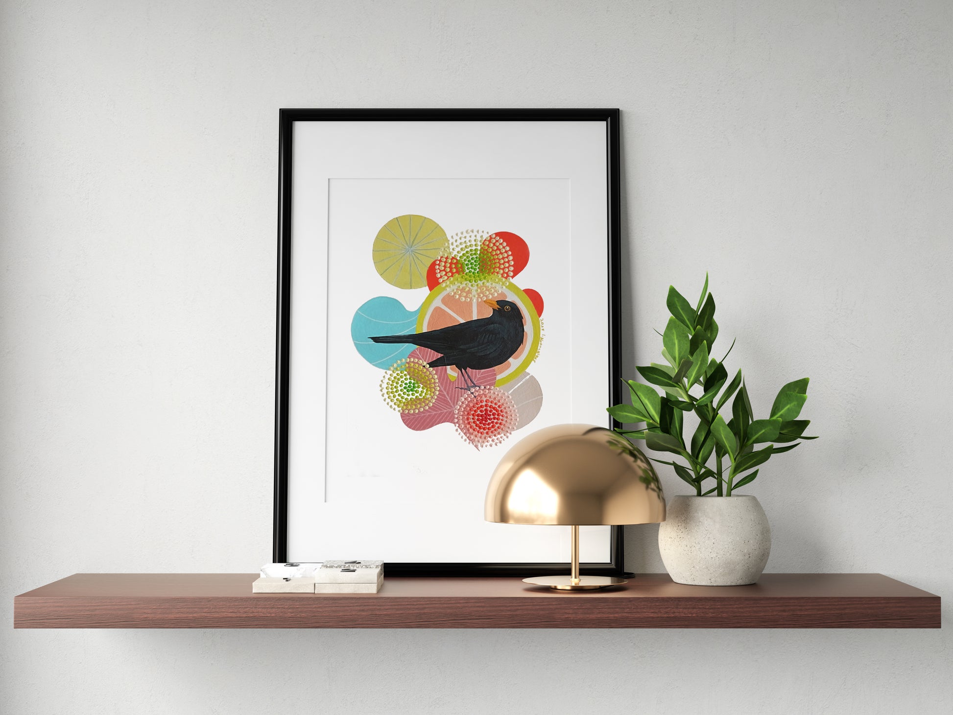 A framed fine art print, by Lis Gonçalves. Open edition, signed by the artist. Printed in Hahnemühle matt fibre paper; featuring a black bird silhouette over colorful abstract circles, displayed on a wooden shelf next to a gold dome lamp and a potted green plant against a white wall.