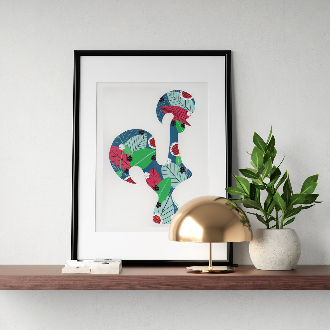 A framed fine art print, by Lis Gonçalves. Open edition, signed by the artist (printed in Hahnemühle matt fibre paper) featuring a colorful Portuguese rooster in a colorful tropical floral pattern, displayed on a wooden shelf next to a gold dome lamp and a potted green plant against a light gray wall.