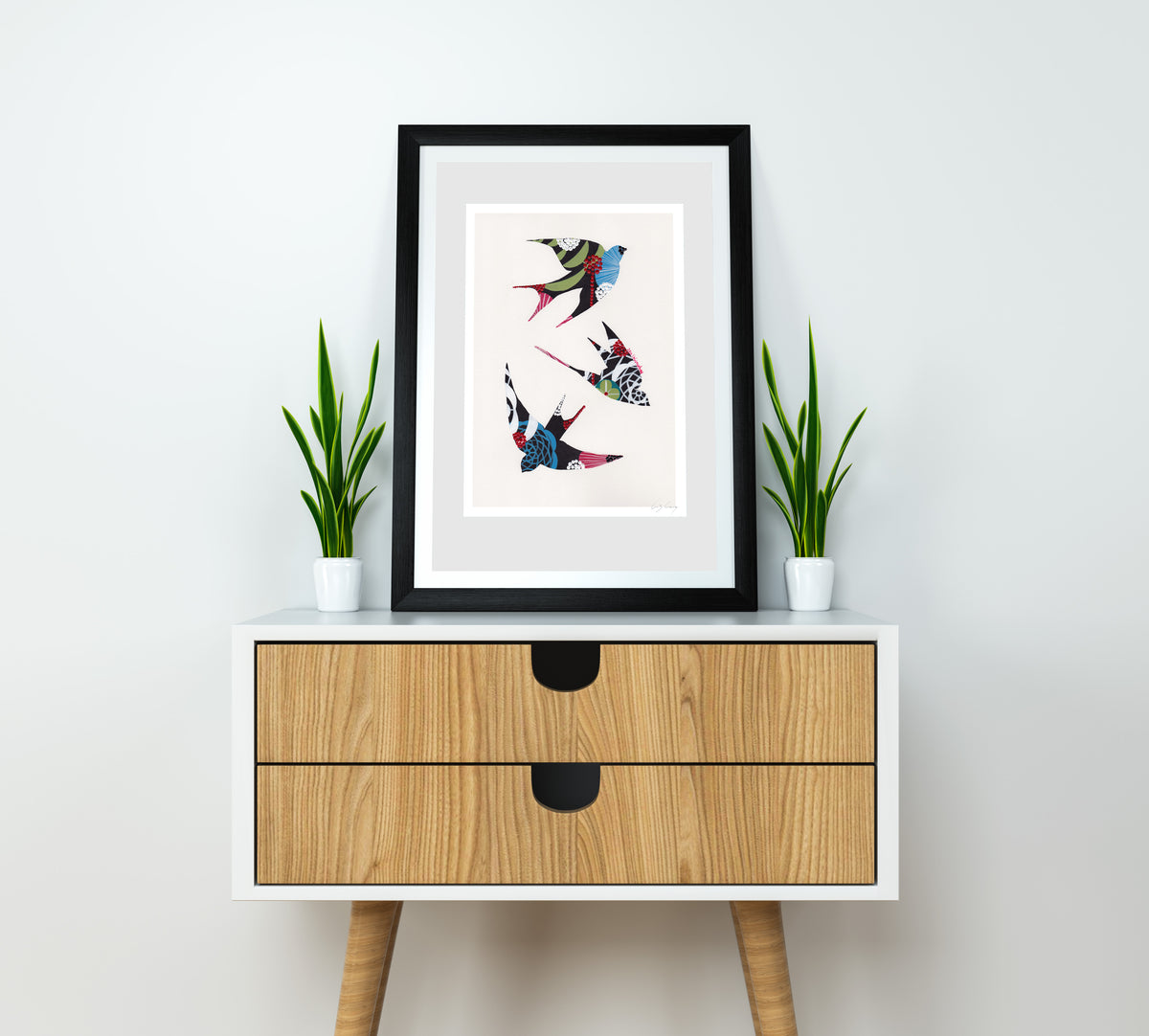 A framed fine art print, by Lis Gonçalves. Open edition, signed by the artist (printed in Hahnemühle matt fibre paper) featuring colorful swallows on a white wall above a wooden cabinet with plants.