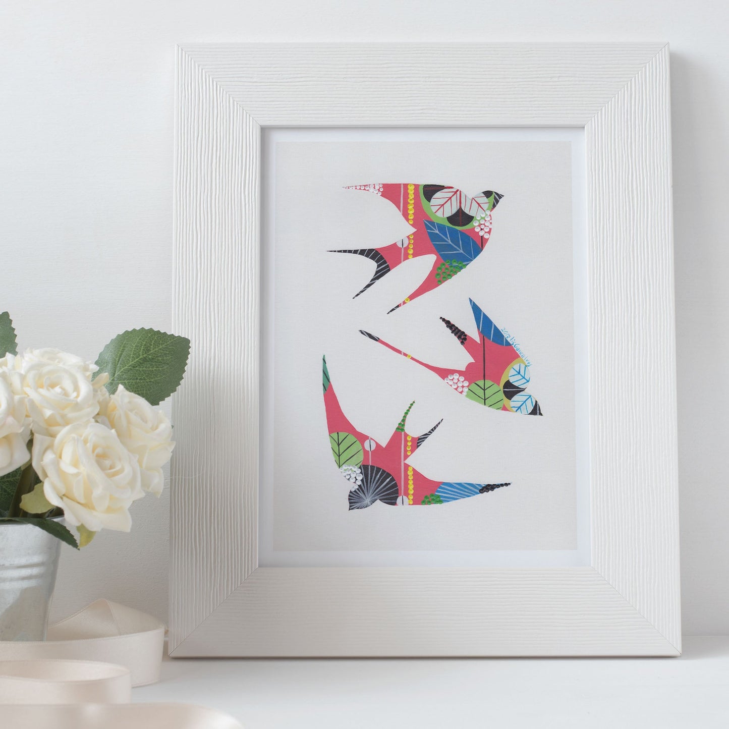 A framed fine art print, by Lis Gonçalves. Open edition, signed by the artist (printed in Hahnemühle matt fibre paper) featuring colorful swallows next to a vase of white flowers on a white surface.