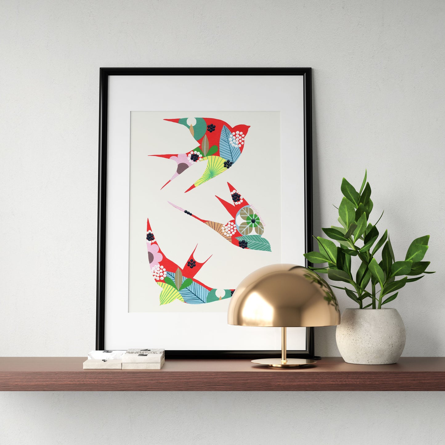 An original artwork by Lis Gonçalves. Painted in acrylic on paper, featuring colorful swallows on a white background, displayed on a floating wooden shelf next to a gold dome lamp and a potted green plant against a light gray wall.
