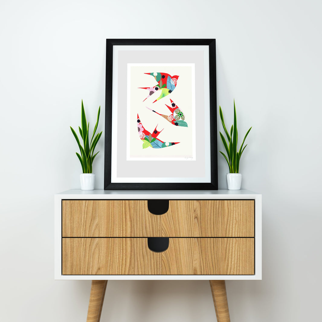 A framed fine art print, by Lis Gonçalves. Open edition, signed by the artist (printed in Hahnemühle matt fibre paper) featuring colorful swallows, styled on a white and wood sideboard with two potted green plants against a white wall.