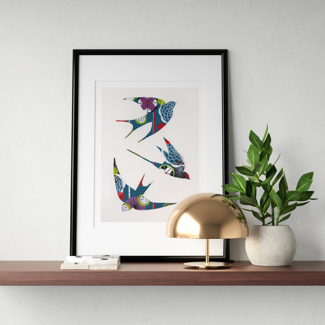 A framed fine art print, by Lis Gonçalves. Open edition, signed by the artist (printed in Hahnemühle matt fibre paper) featuring colorful swallows, styled on a wooden shelf next to a gold dome lamp and a potted green plant against a light gray wall.