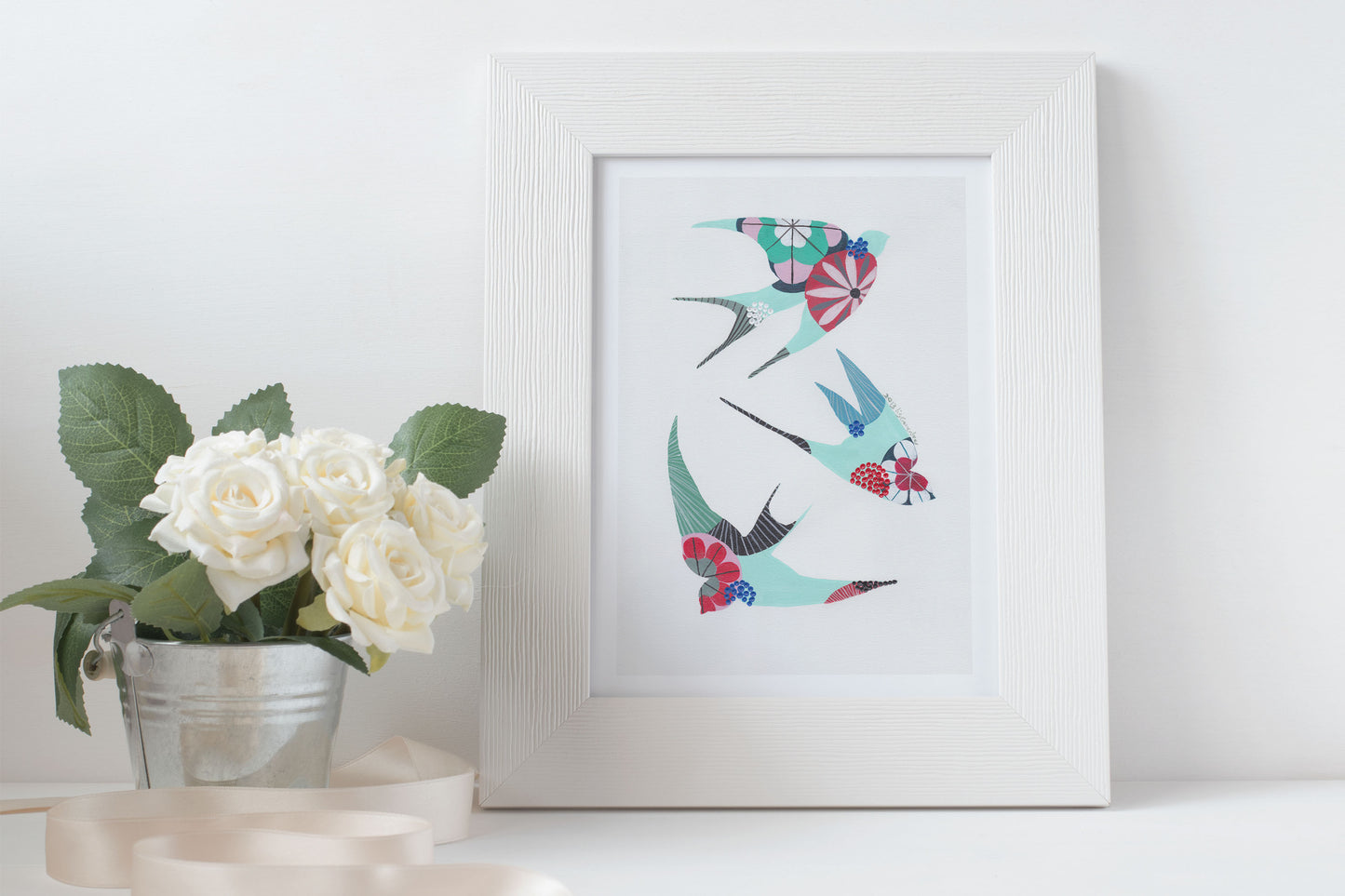 A framed fine art print, by Lis Gonçalves. Open edition, signed by the artist (printed in Hahnemühle matt fibre paper) featuring colorful swallows on a white surface next to a small potted plant.