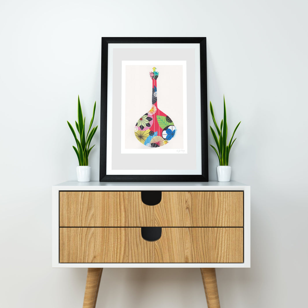 A framed fine art print, by Lis Gonçalves. Open edition, signed by the artist (printed in Hahnemühle matt fibre paper) featuring a colorful Portuguese guitar, styled on a white and wood sideboard with two potted green plants against a white wall.
