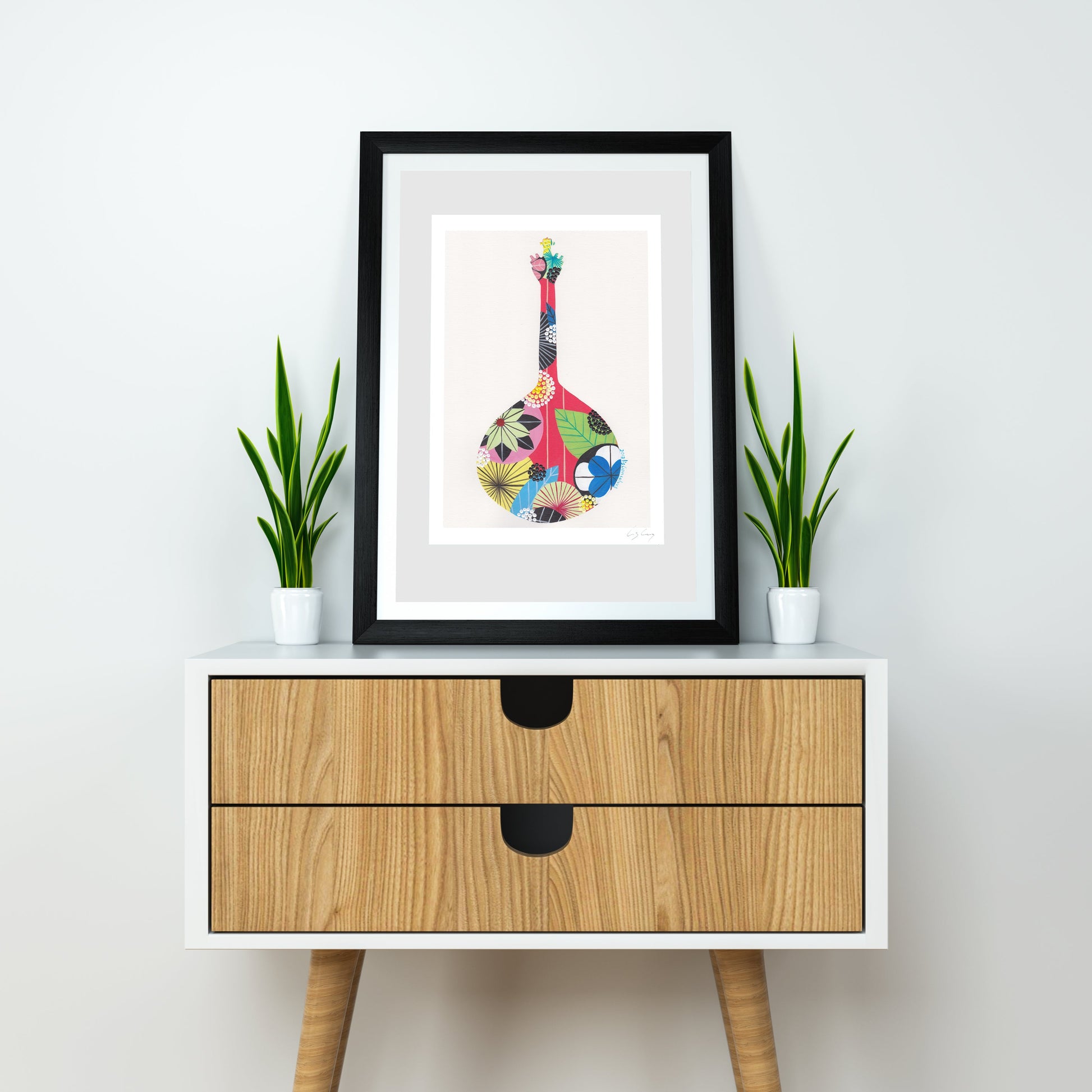 A framed fine art print, by Lis Gonçalves. Open edition, signed by the artist (printed in Hahnemühle matt fibre paper) featuring a colorful Portuguese guitar, styled on a white and wood sideboard with two potted green plants against a white wall.