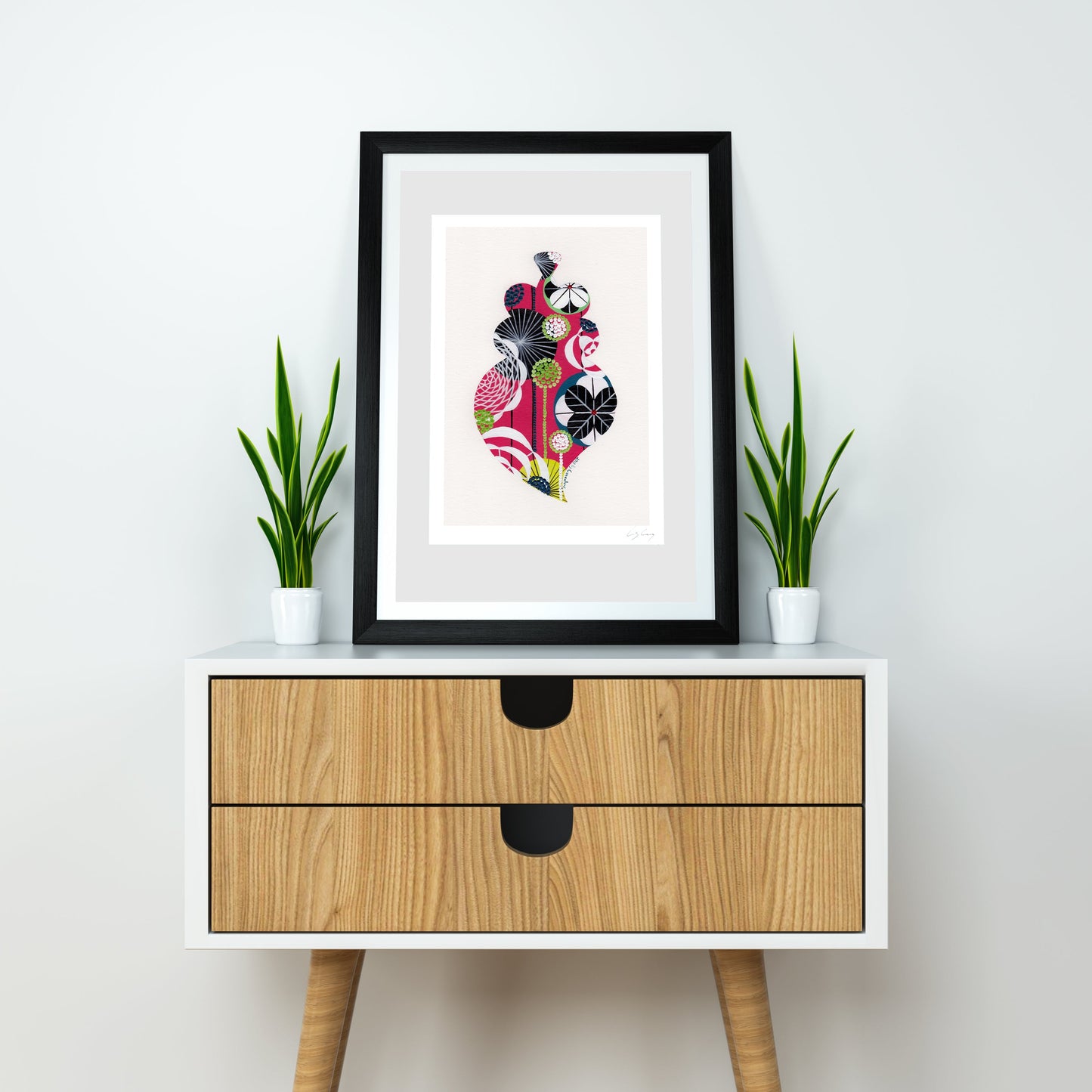 A framed fine art print, by Lis Gonçalves. Open edition, signed by the artist (printed in Hahnemühle matt fibre paper) featuring a colorful Portuguese Viana heart on a white and wood mid-century modern sideboard with two potted green plants against a white wall.
