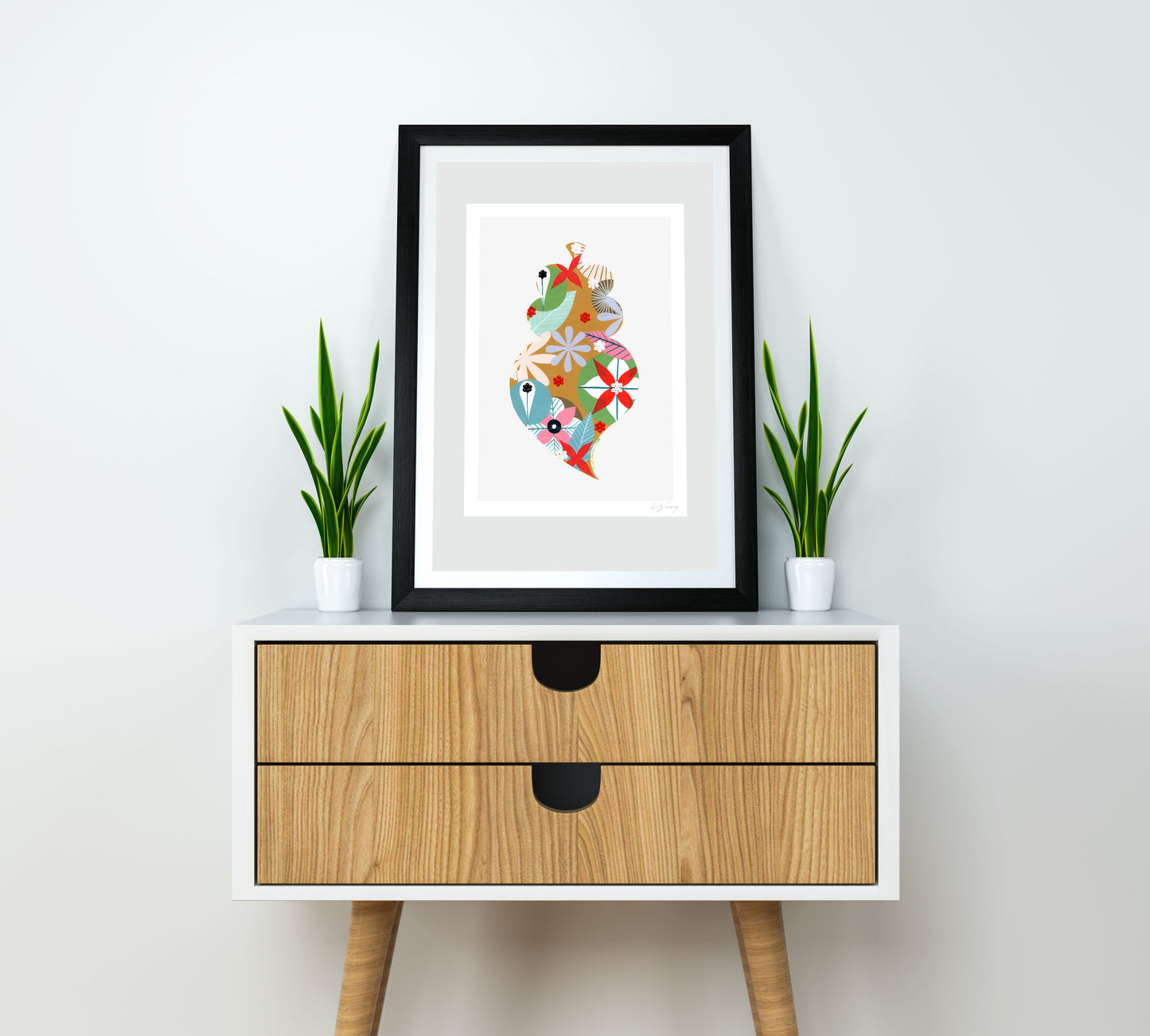 A framed fine art print, by Lis Gonçalves. Open edition, signed by the artist (printed in Hahnemühle matt fibre paper) featuring a colorful Portuguese Viana heart on a white wall above a wooden cabinet with plants.