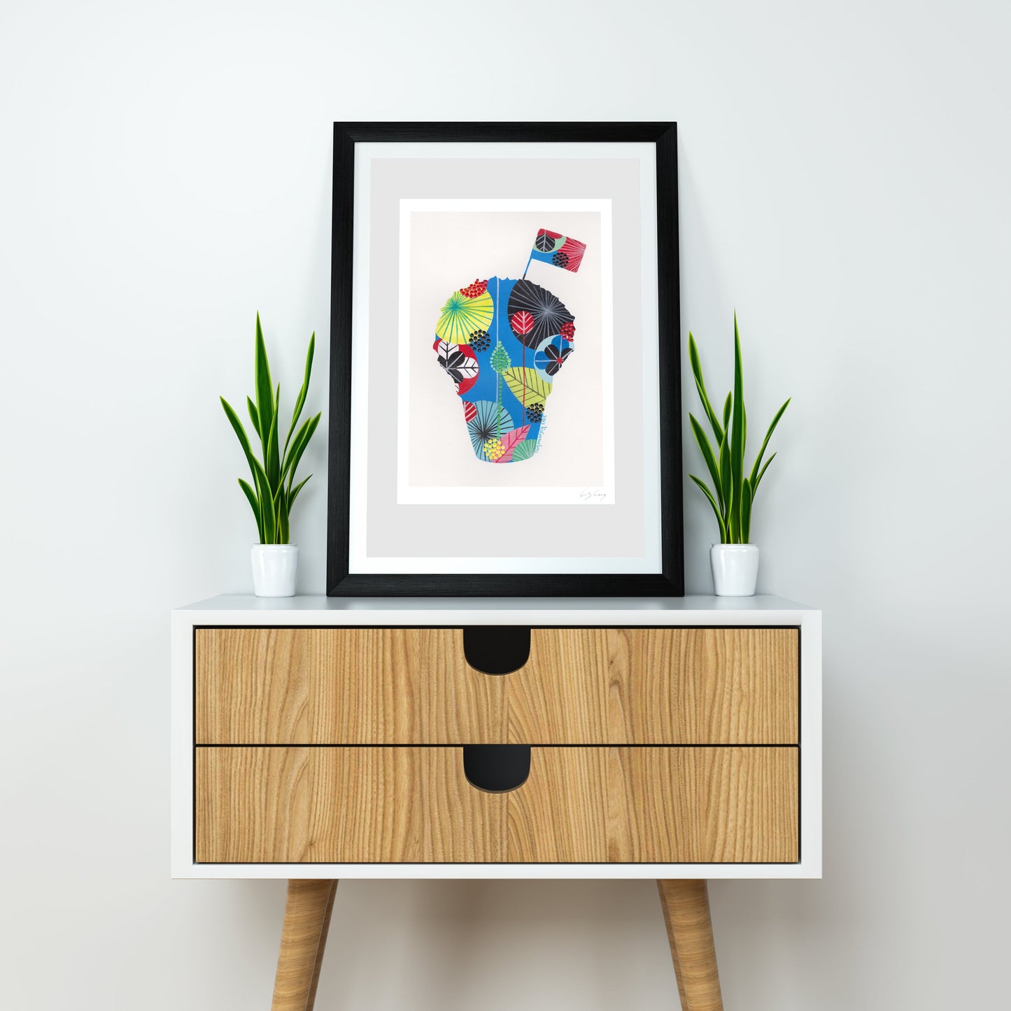 A framed fine art print, by Lis Gonçalves. Open edition, signed by the artist (printed in Hahnemühle matt fibre paper) featuring a colorful, patterned silhouette of a manjerico basil plant, styled on a light wood and white two-drawer sideboard with two potted green plants against a white wall.