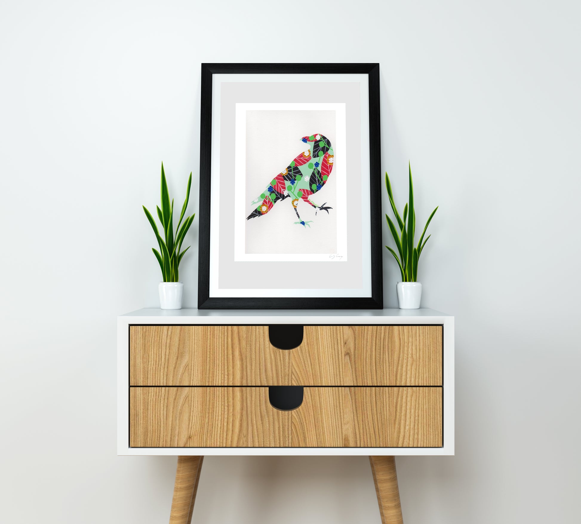 A framed fine art print, by Lis Gonçalves. Open edition, signed by the artist (printed in Hahnemühle matt fibre paper) featuring a colorful raven on a white wall above a wooden cabinet with two drawers.