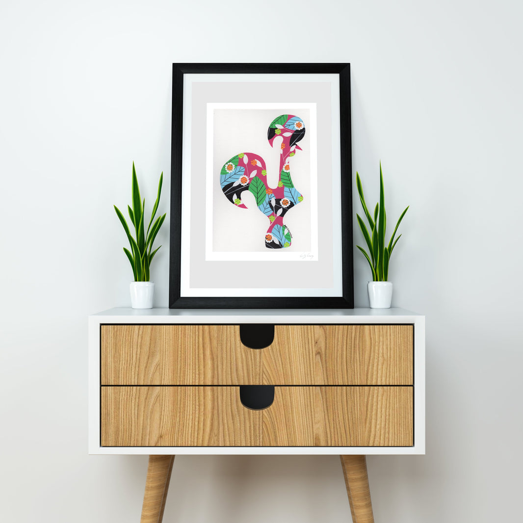 A framed fine art print, by Lis Gonçalves. Open edition, signed by the artist (printed in Hahnemühle matt fibre paper) featuring a colorful Portuguese rooster, displayed on a white sideboard with two small potted green plants on either side. The sideboard has a light wood grain finish and black drawer pulls, set against a plain white wall.