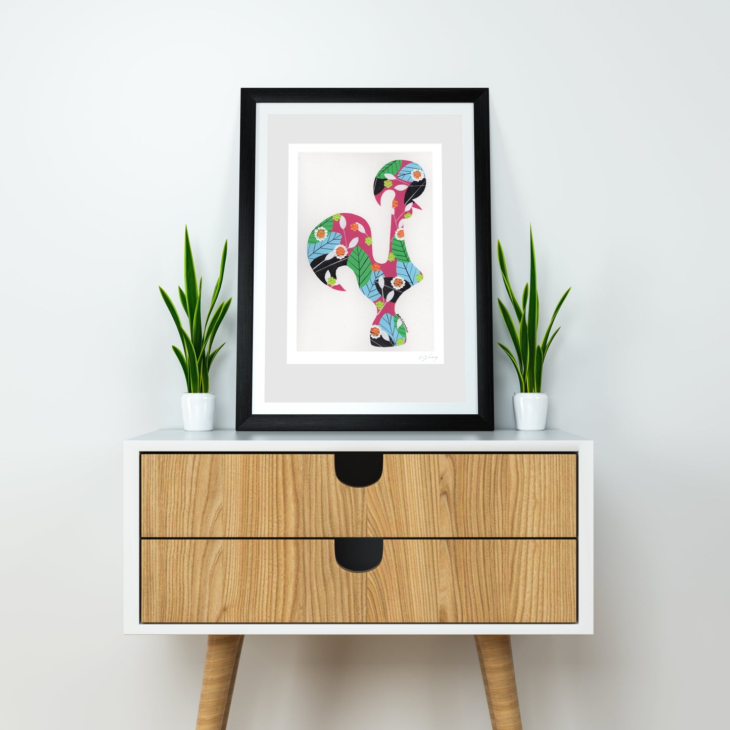 A framed fine art print, by Lis Gonçalves. Open edition, signed by the artist (printed in Hahnemühle matt fibre paper) featuring a colorful Portuguese rooster, displayed on a white sideboard with two small potted green plants on either side. The sideboard has a light wood grain finish and black drawer pulls, set against a plain white wall.