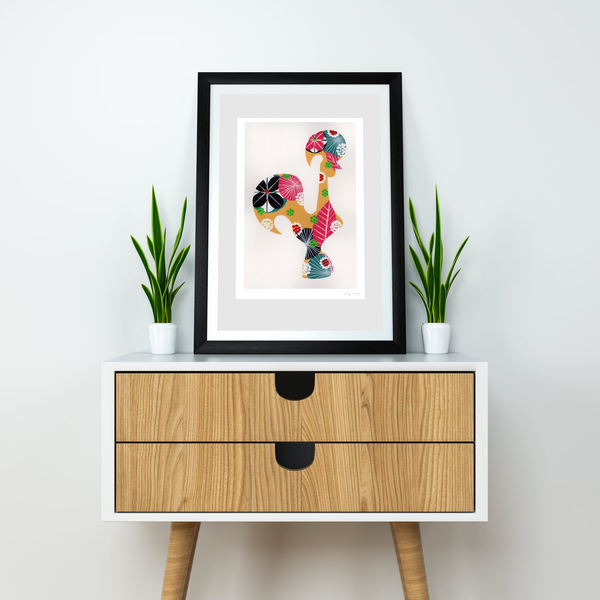 A framed fine art print, by Lis Gonçalves. Open edition, signed by the artist (printed in Hahnemühle matt fibre paper) featuring a colorful Portuguese rooster , styled on a white sideboard with two small potted green plants on either side.