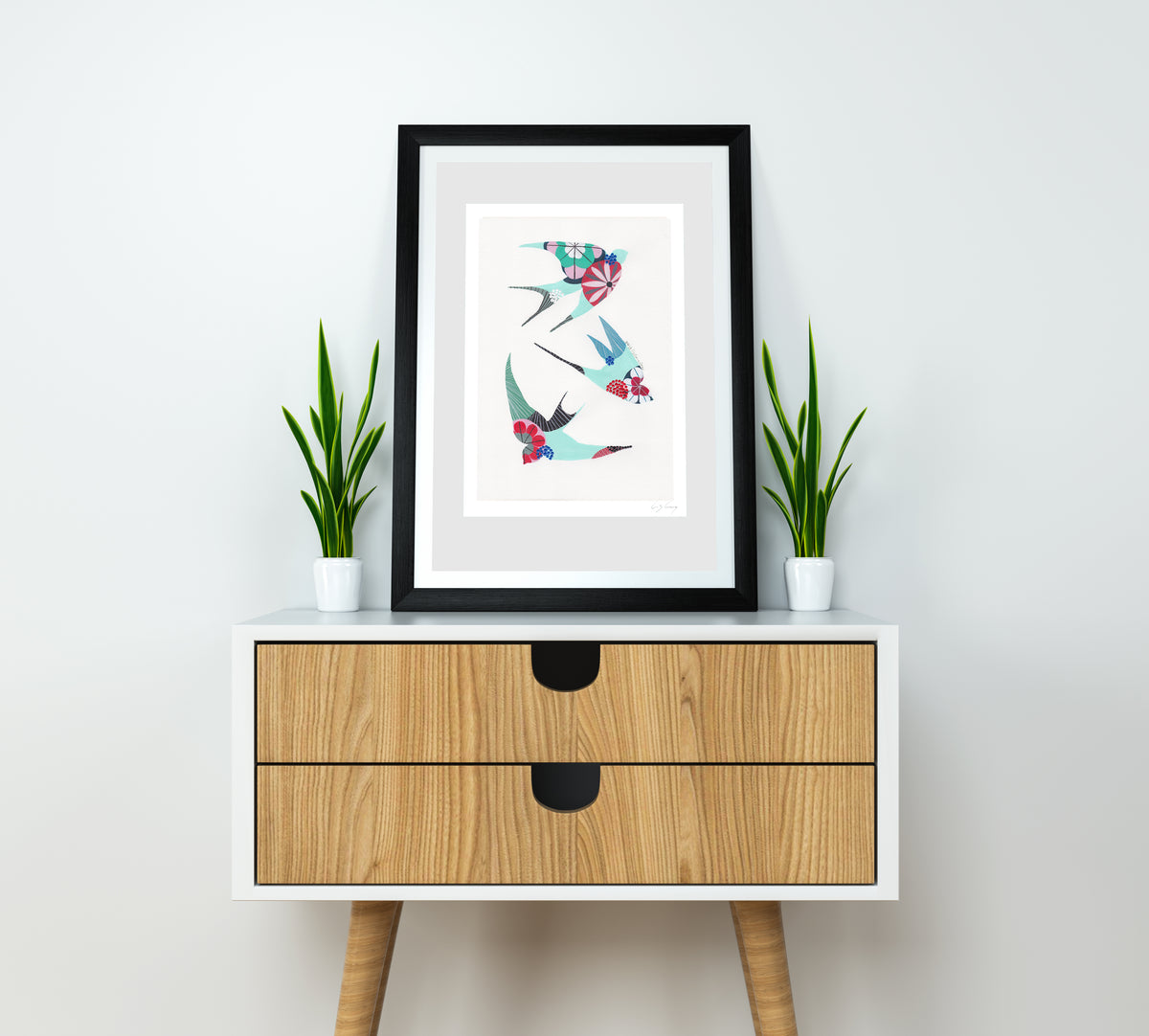 A framed fine art print, by Lis Gonçalves. Open edition, signed by the artist (printed in Hahnemühle matt fibre paper) featuring colorful swallows on a white wall above a wooden cabinet with two drawers.
