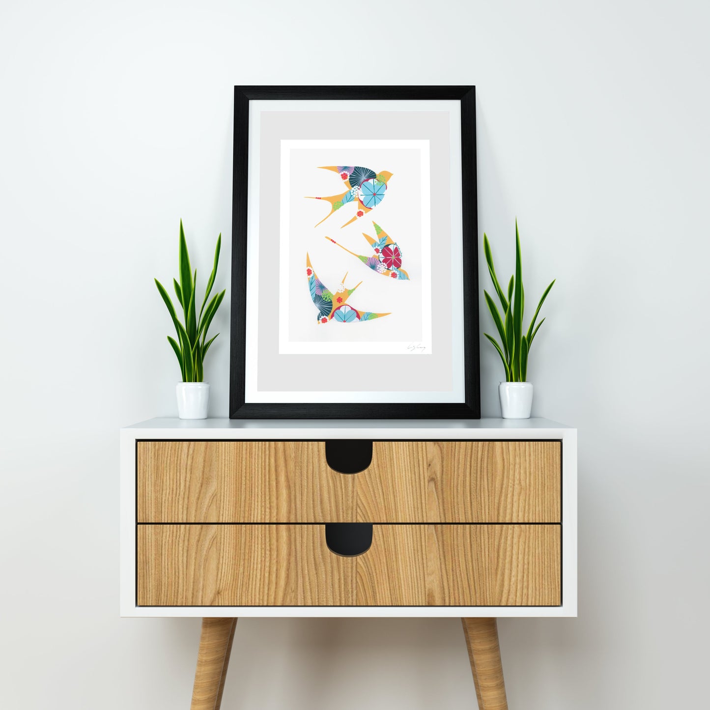 A framed fine art print, by Lis Gonçalves. Open edition, signed by the artist (printed in Hahnemühle matt fibre paper) featuring colorful swallows on a white wall above a wooden cabinet with plants.