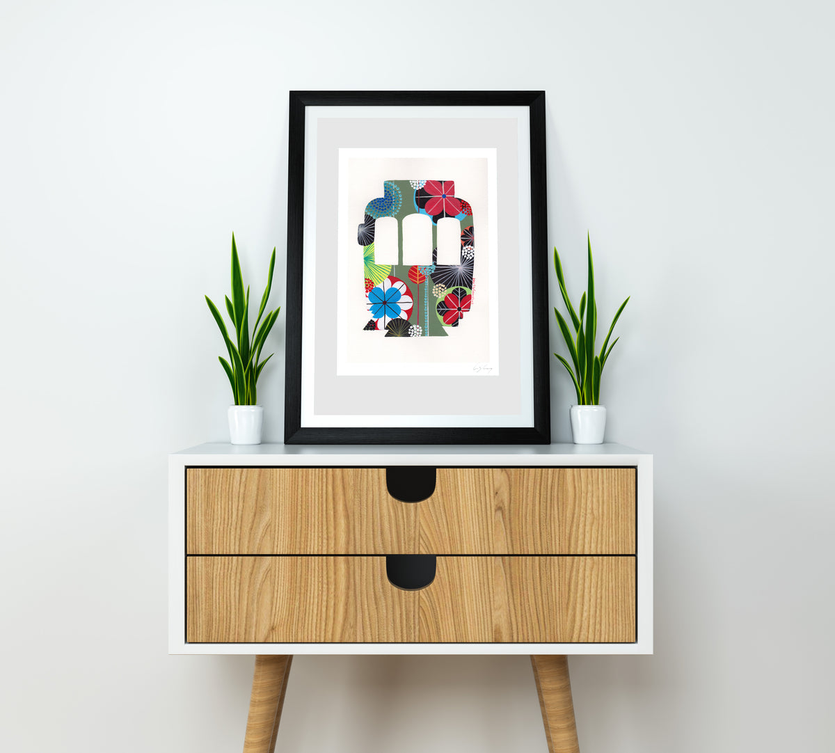 A framed fine art print, by Lis Gonçalves. Open edition, signed by the artist (printed in Hahnemühle matt fibre paper) featuring a colorful Lisbon tram on a white wall above a wooden cabinet.