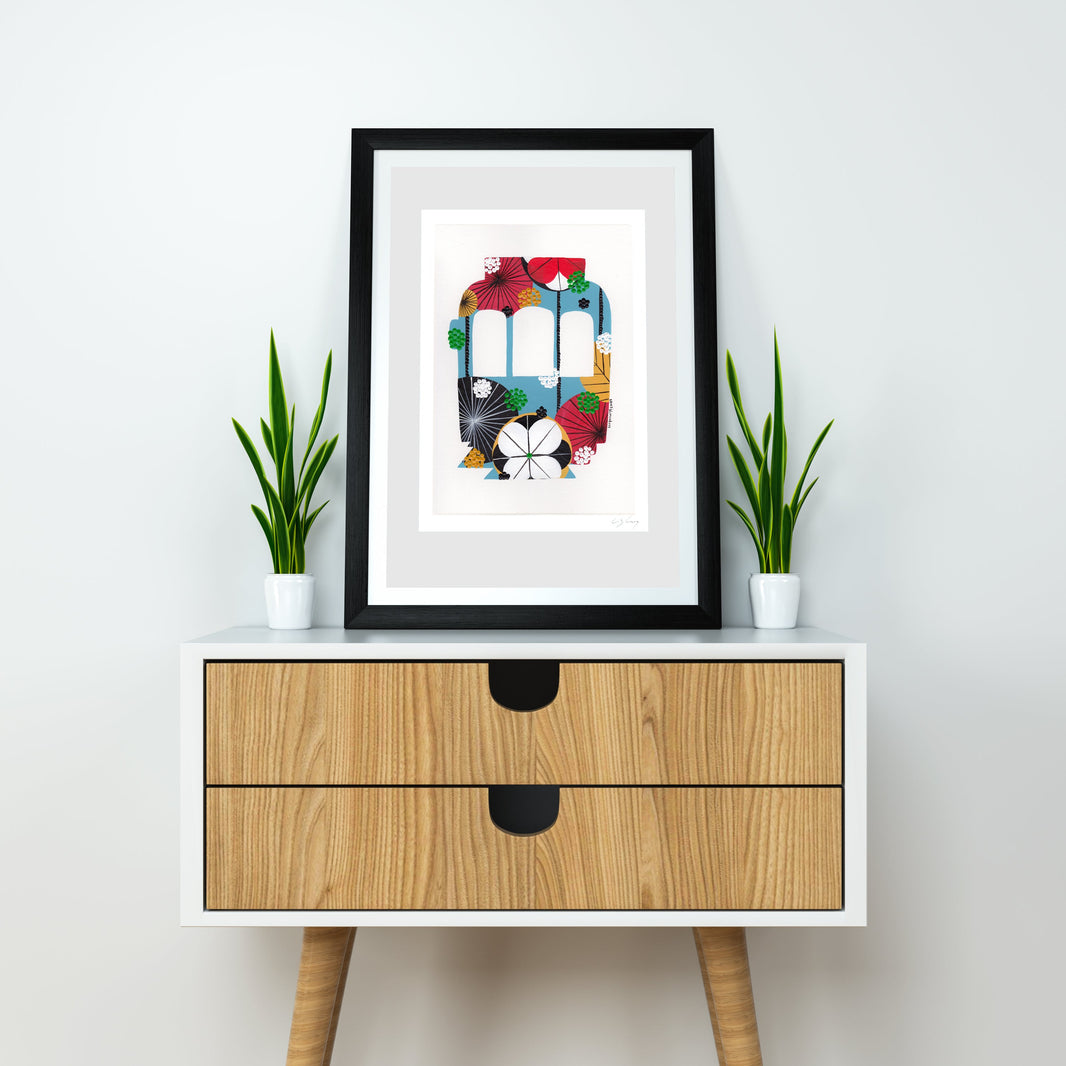 A framed fine art print, by Lis Gonçalves. Open edition, signed by the artist (printed in Hahnemühle matt fibre paper) featuring a colorful Lisbon tram, styled on a white cabinet with two small potted green plants against a light gray wall.