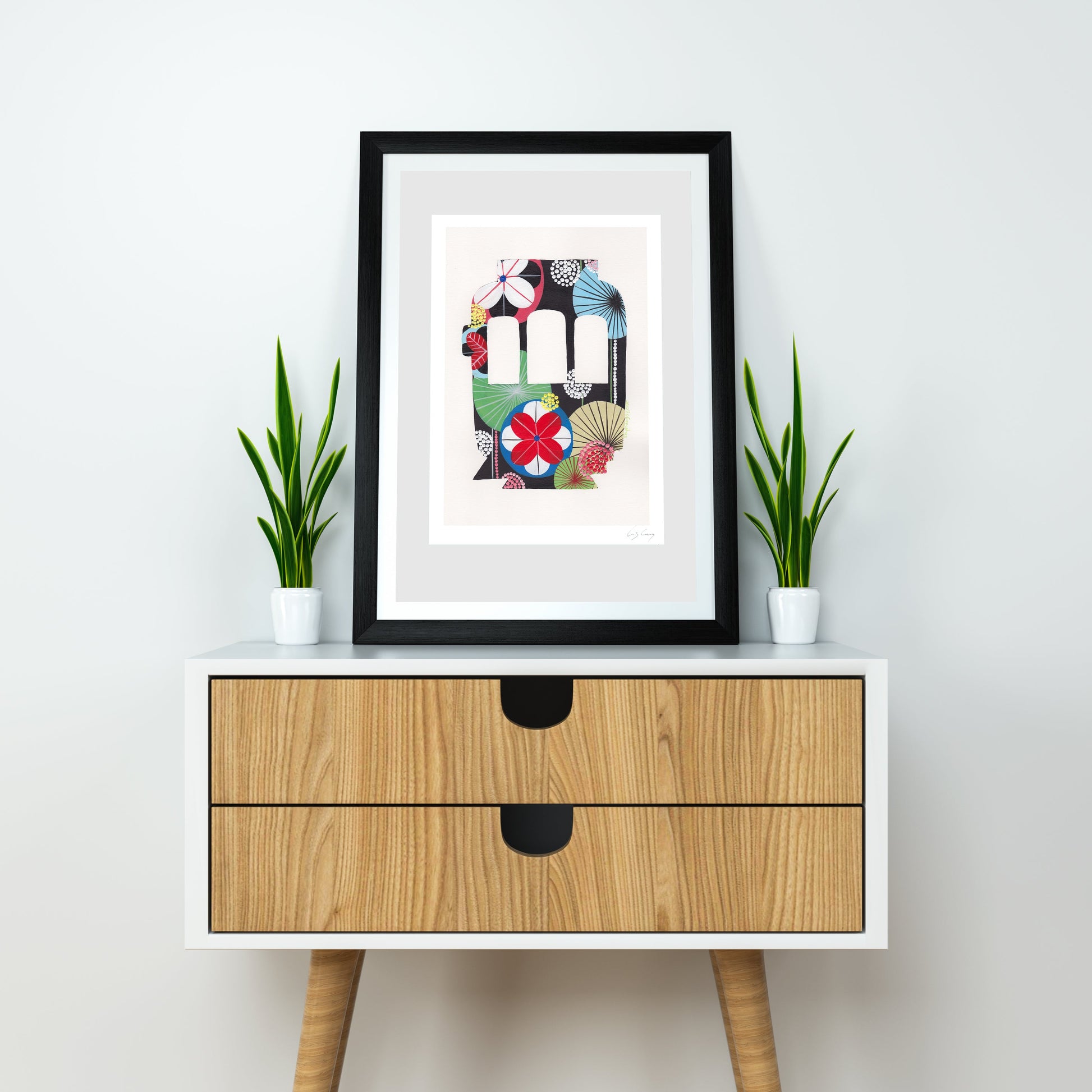 A framed fine art print, by Lis Gonçalves. Open edition, signed by the artist (printed in Hahnemühle matt fibre paper) featuring a colorful Lisbon tram, displayed on a white and wood sideboard with two potted green plants against a light gray wall.