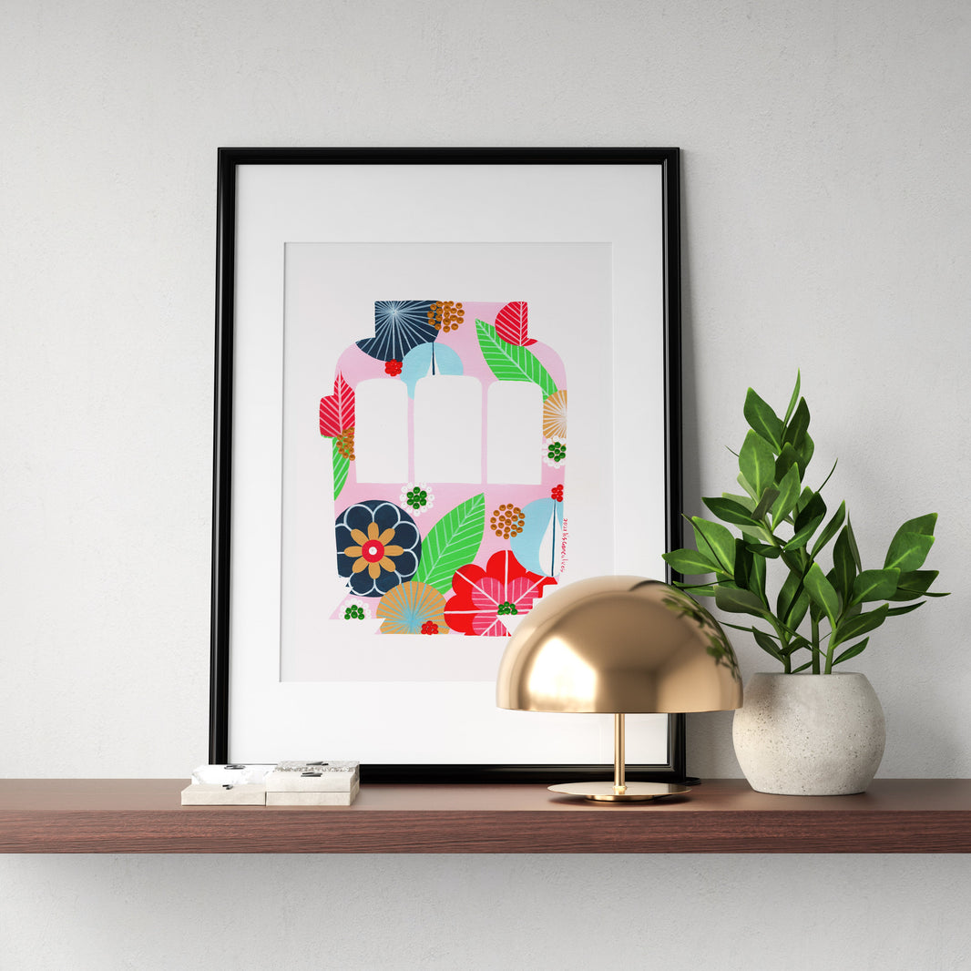 A framed fine art print, by Lis Gonçalves. Open edition, signed by the artist (printed in Hahnemühle matt fibre paper) featuring a colorful Lisbon tram on a wooden shelf with a lamp and plant against a white wall