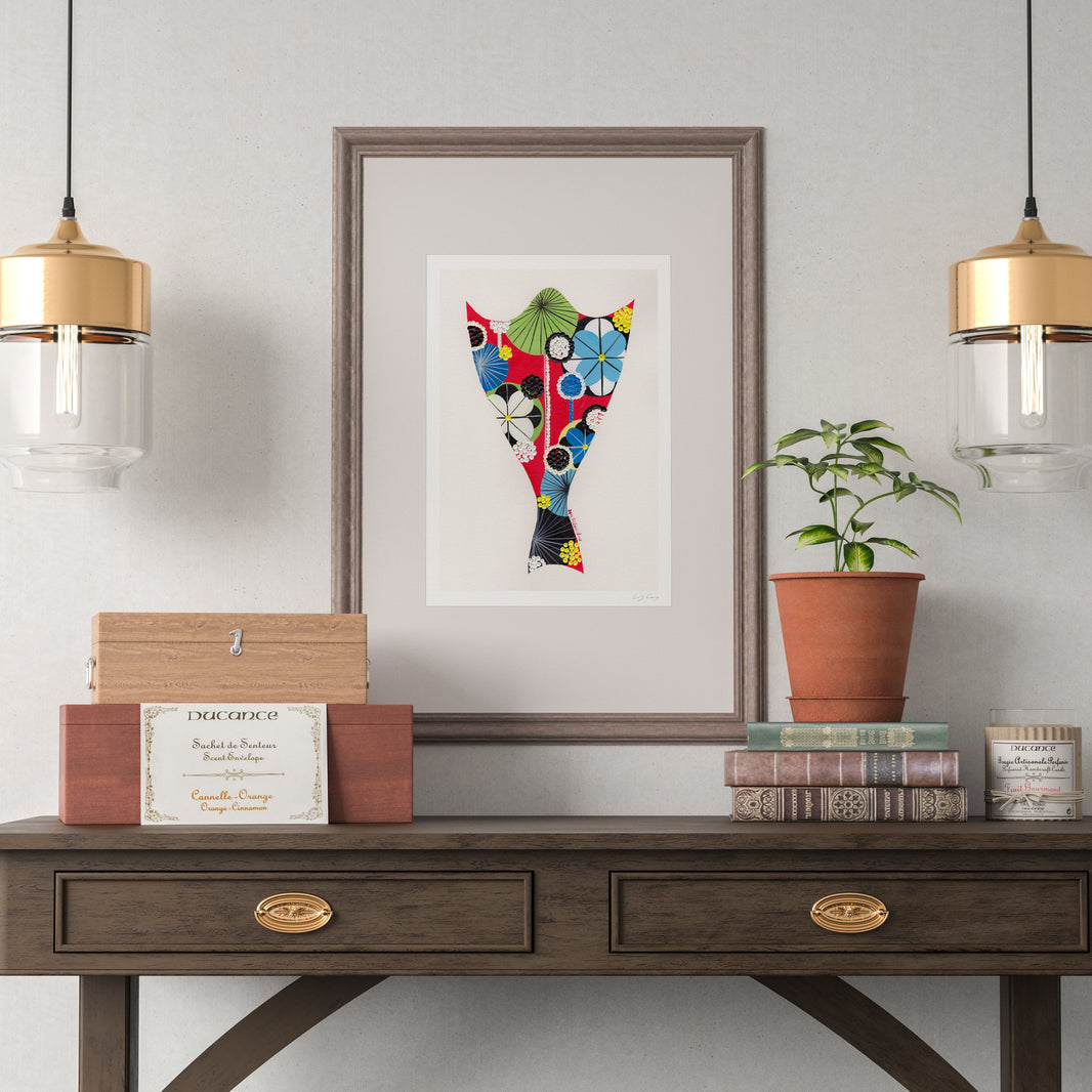 A framed fine art print, by Lis Gonçalves. Open edition, signed by the artist (printed in Hahnemühle matt fibre paper) featuring a colorful, patterned cod fish illustration hangs on a light gray wall above a dark wood console table. The scene is styled with two hanging pendant lights, a small potted plant, books, and decorative boxes.